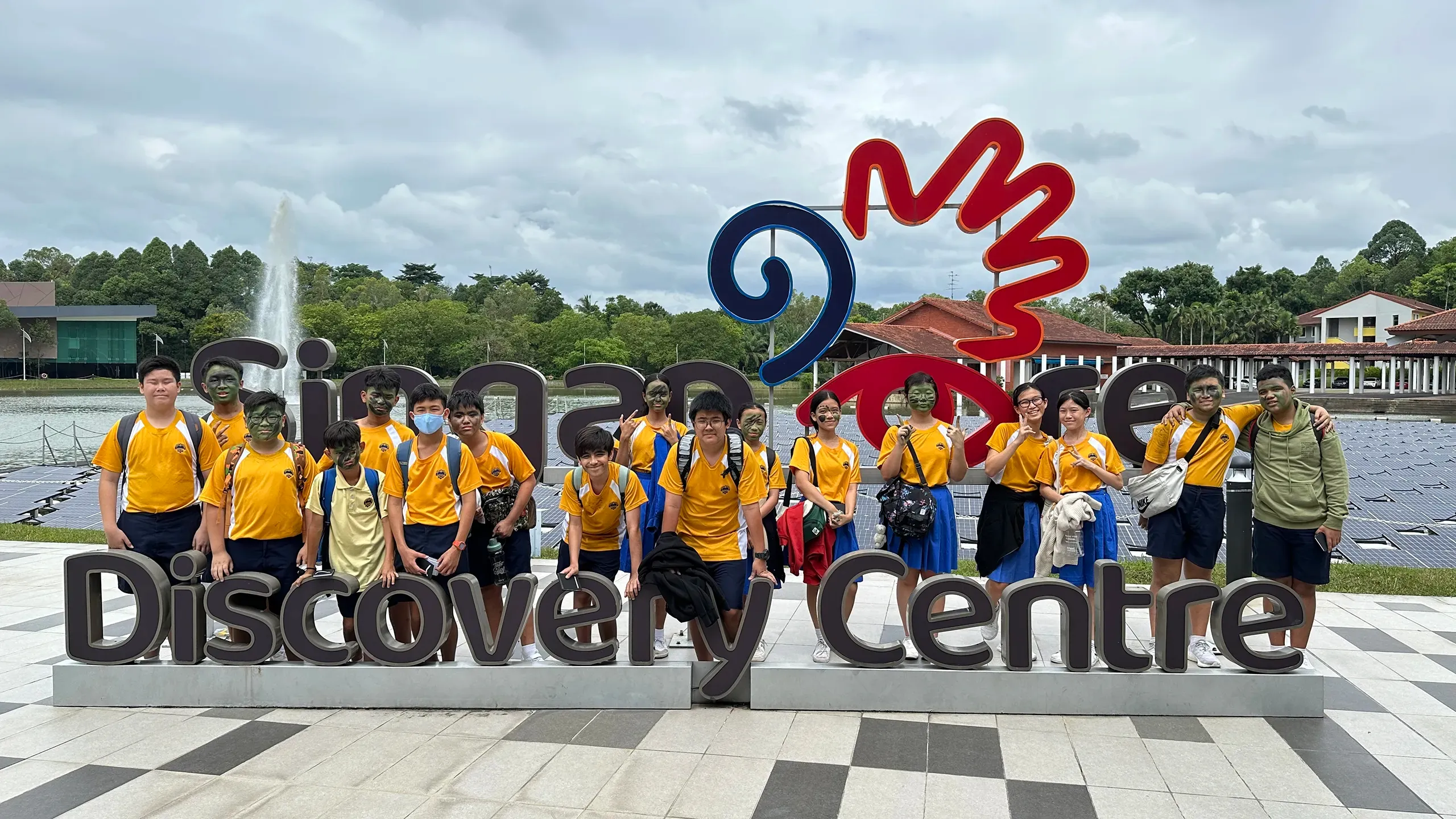 A large group of Secondary 1 students posing together behind a large outdoor sign that reads "Singapore Discovery Centre". The students are wearing their yellow and white school PE shirts paired with bright blue skirts or dark shorts. Many of them are sporting green camouflage face paint from their outdoor survival activities, with a lake, fountain, and floating solar panels visible under a cloudy sky in the background.